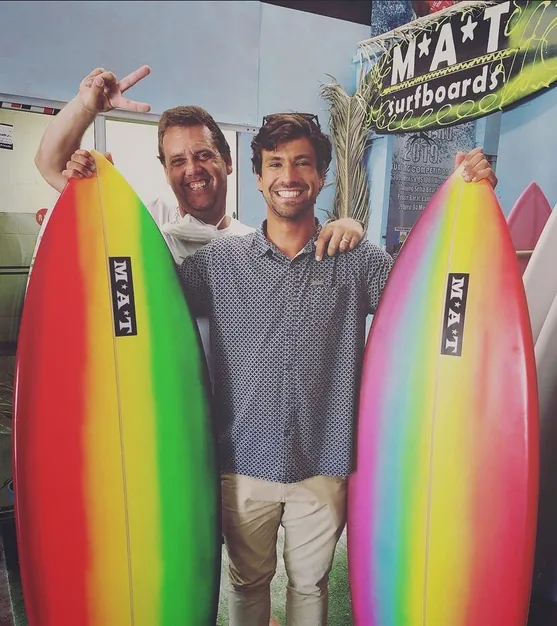Two smiling surfers holding rainbow airbrushed MAT surfboards inside the MAT Surfboards shop